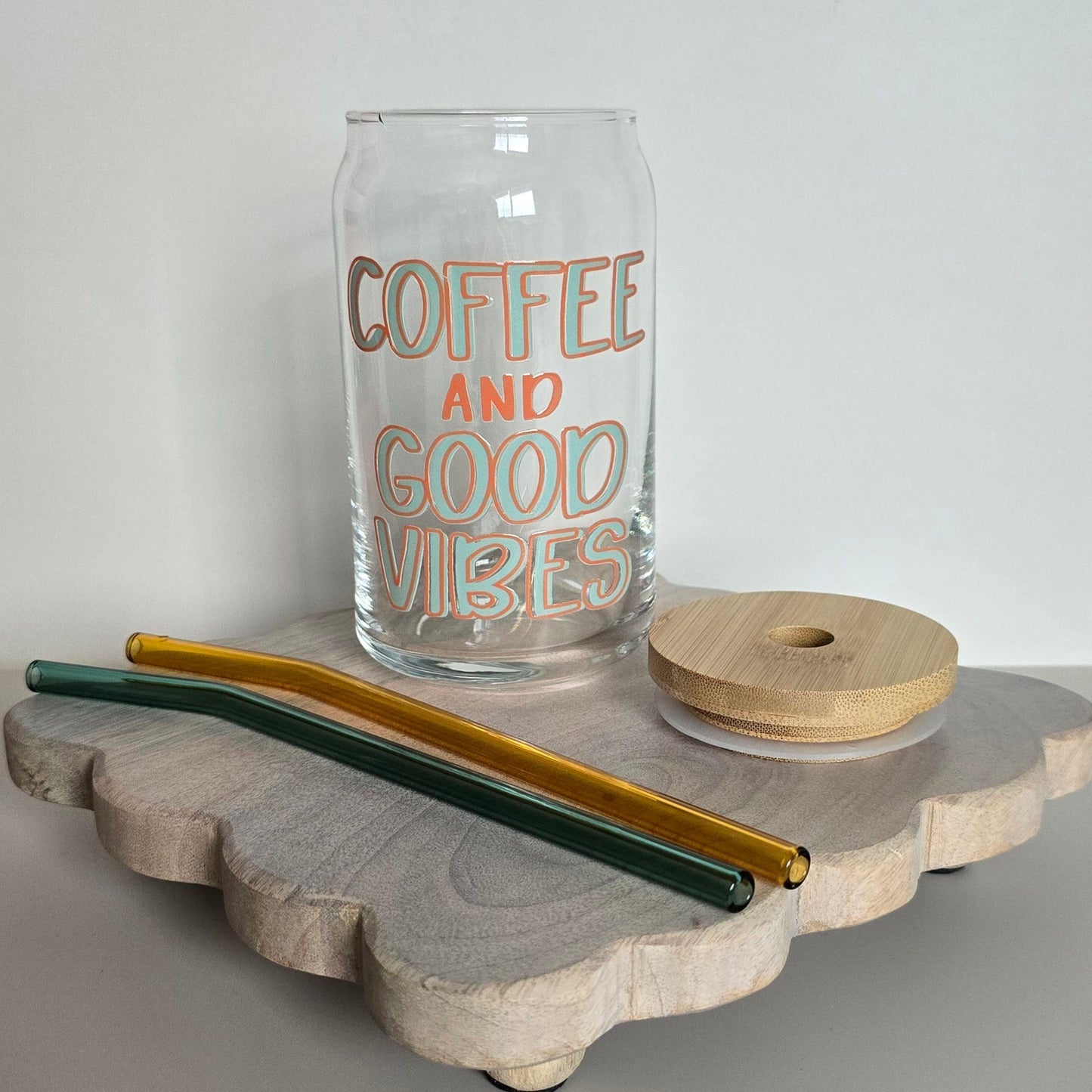 A glass coffee mug with the words 'COFFEE AND GOOD VIBES' printed on it, displayed on a wooden trivet, accompanied by two metal straws with a green and yellow design.